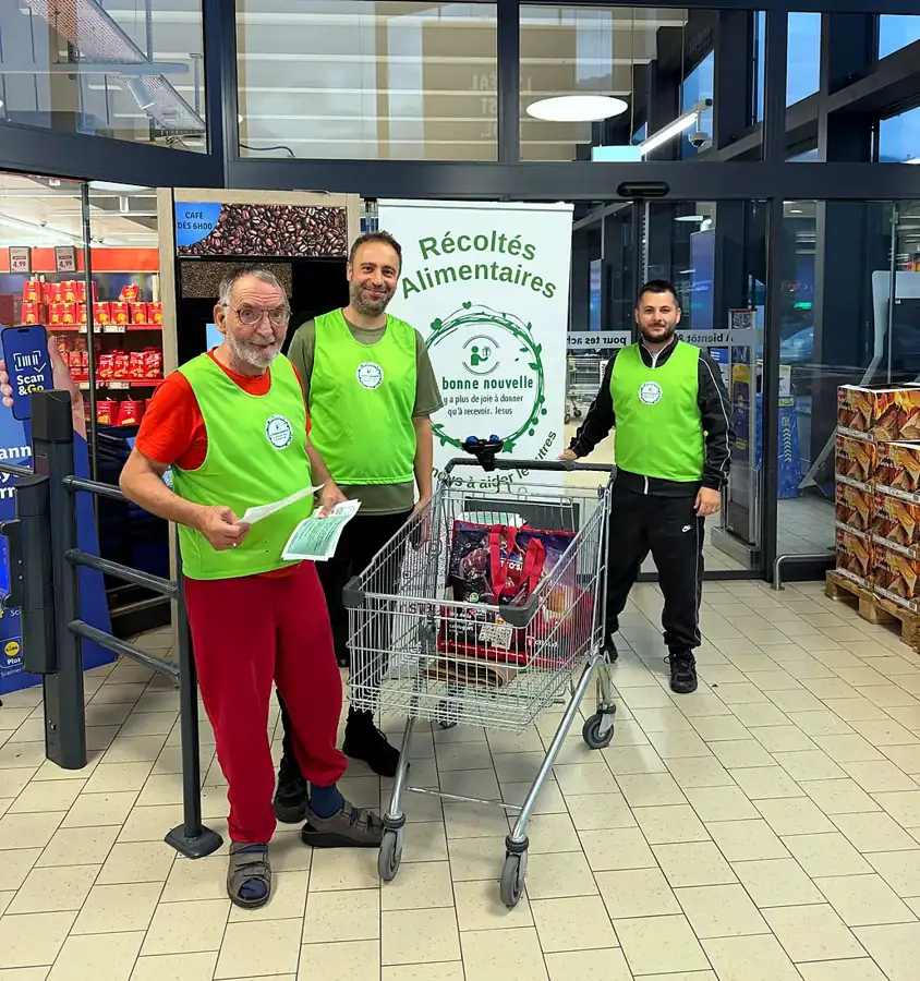 Trois bénévoles en gilet vert avec un chariot de dons devant un magasin